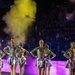 A group of female performers are executing a synchronized jump during a large-scale nighttime performance or halftime show. They are wearing matching metallic gold-and-black patterned bodysuits with silver skirts and silver lace-up heels. The venue appears to be a stadium filled with spectators, with dramatic yellow smoke and pink/purple lighting effects enhancing the spectacle.