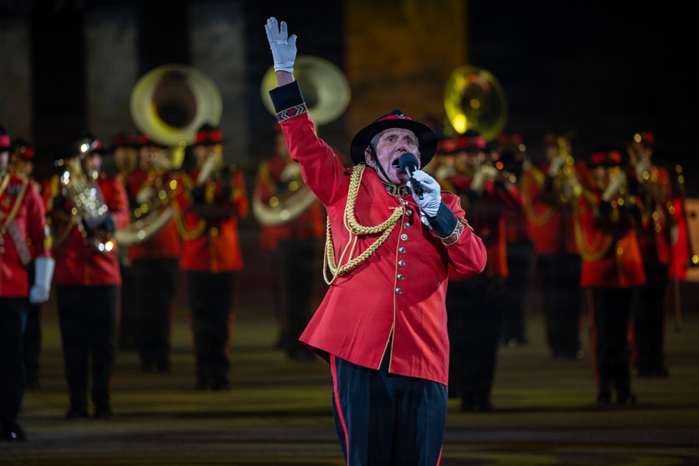 A military band performance taking place at night, featuring a prominent announcer or master of ceremonies in the foreground dressed in a red ceremonial uniform with gold braiding. He is holding a microphone and raising one white-gloved hand, addressing the audience. Behind him, a large brass band in matching red uniforms performs, with visible tubas and other brass instruments.