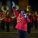 A military band performance taking place at night, featuring a prominent announcer or master of ceremonies in the foreground dressed in a red ceremonial uniform with gold braiding. He is holding a microphone and raising one white-gloved hand, addressing the audience. Behind him, a large brass band in matching red uniforms performs, with visible tubas and other brass instruments.