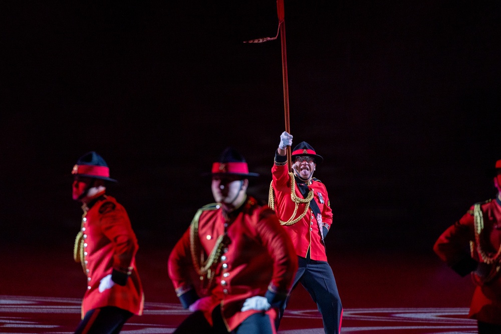 A group of performers dressed in red ceremonial uniforms with black hats and gold braiding are performing in a darkened arena or stadium. The central figure is prominently in focus, holding a long staff or lance raised high while making an expressive face. The uniforms closely resemble those of the Royal Canadian Mounted Police (RCMP), suggesting this may be a ceremonial musical ride or tattoo performance.