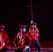 A group of performers dressed in red ceremonial uniforms with black hats and gold braiding are performing in a darkened arena or stadium. The central figure is prominently in focus, holding a long staff or lance raised high while making an expressive face. The uniforms closely resemble those of the Royal Canadian Mounted Police (RCMP), suggesting this may be a ceremonial musical ride or tattoo performance.