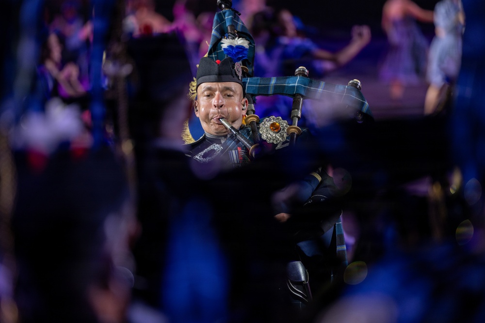 A bagpiper in traditional Scottish military dress uniform is performing at what appears to be a large ceremonial event or tattoo. The musician is captured mid-performance, playing the bagpipes with eyes closed and cheeks puffed. The image is shot with a shallow depth of field, with blurred figures in the foreground and background suggesting a large ensemble performance.