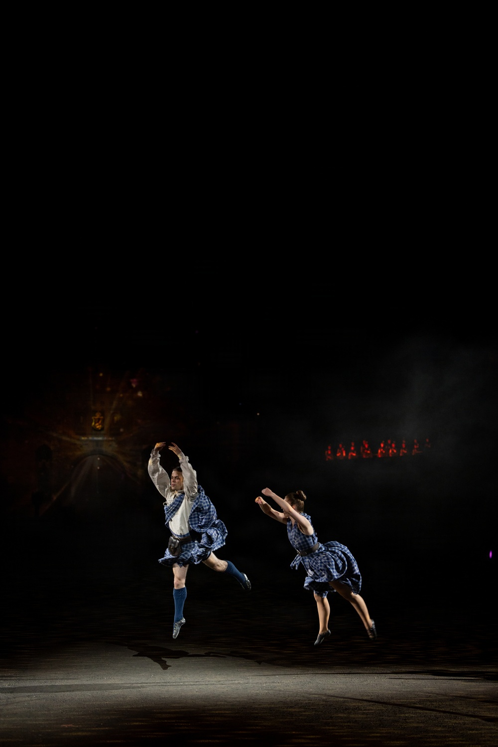 Two dancers are captured mid-air performing what appears to be Scottish Highland dancing on a darkened stage. Both wear matching blue and white plaid/tartan costumes with short skirts, and one dancer is also wearing blue knee-high socks. The dramatic low-key lighting isolates the performers against a nearly black background, with faint red stage lights visible in the distance.