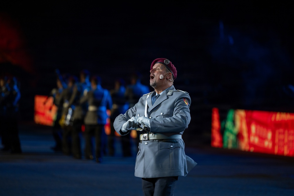 A military performer in a gray uniform and burgundy/maroon beret is singing or performing at what appears to be a formal military ceremony or tattoo event. He wears a wireless microphone on his cheek and white gloves, with his hands clasped in front of him. In the background, other uniformed personnel are visible under dramatic blue stage lighting with colorful LED displays.