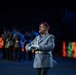 A military performer in a gray uniform and burgundy/maroon beret is singing or performing at what appears to be a formal military ceremony or tattoo event. He wears a wireless microphone on his cheek and white gloves, with his hands clasped in front of him. In the background, other uniformed personnel are visible under dramatic blue stage lighting with colorful LED displays.
