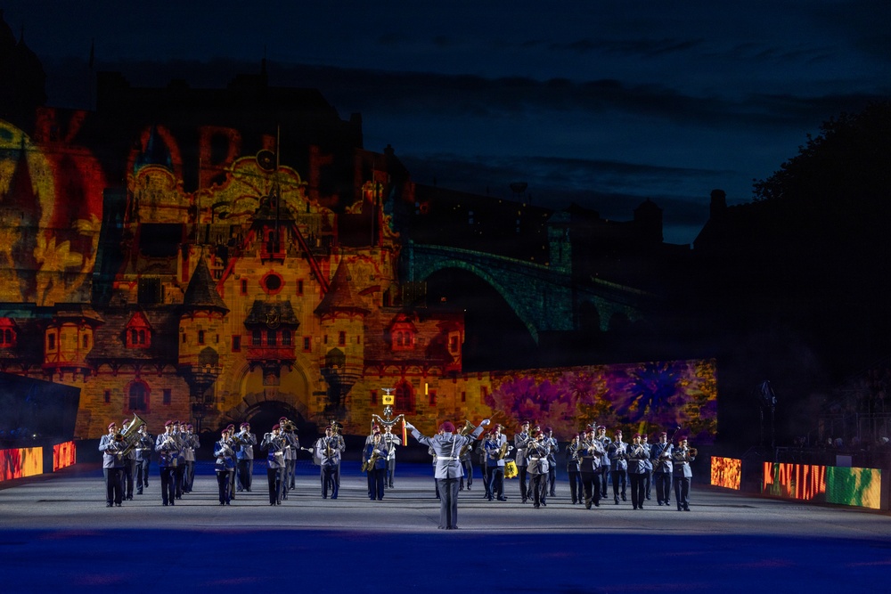 A military marching band performs at night in front of a large stone castle or fortress illuminated by colorful projected light displays. A conductor leads the uniformed musicians across a wide parade ground, with the historic structure serving as a dramatic backdrop. The scene appears to be a formal military tattoo or ceremonial outdoor performance event.