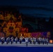 A military marching band performs at night in front of a large stone castle or fortress illuminated by colorful projected light displays. A conductor leads the uniformed musicians across a wide parade ground, with the historic structure serving as a dramatic backdrop. The scene appears to be a formal military tattoo or ceremonial outdoor performance event.