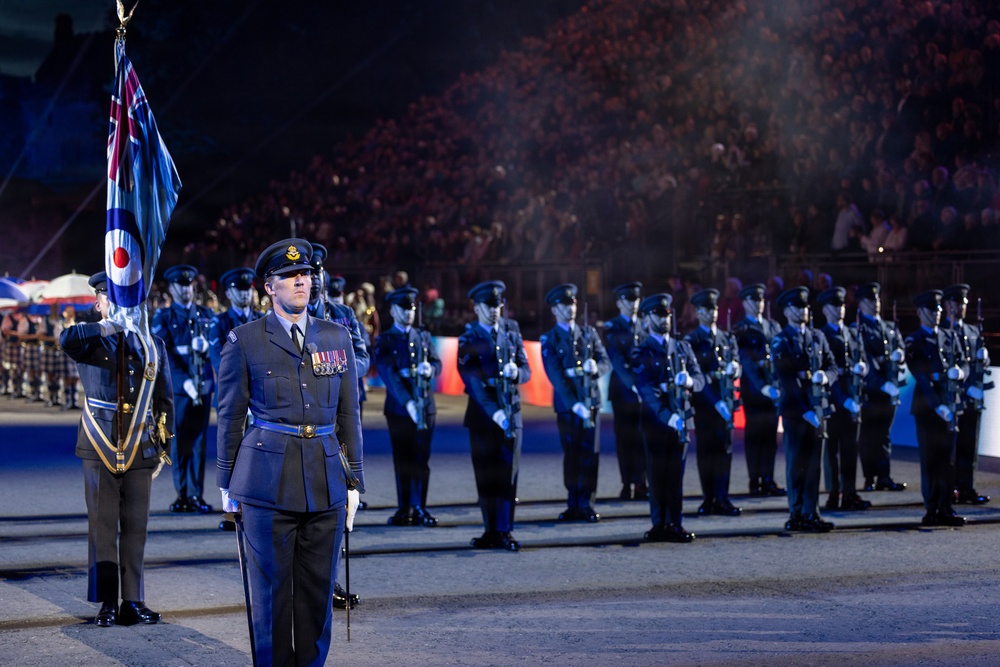 A formal military parade featuring personnel in Royal Air Force (RAF) dress uniforms performing at what appears to be a large nighttime ceremonial event, likely the Royal Edinburgh Military Tattoo or a similar prestigious military pageant. A senior officer stands at the forefront wearing multiple service medals, with a flag bearer displaying the RAF ensign (featuring the Union Jack and RAF roundel) to the left. Rows of uniformed RAF personnel stand at attention in the background before a large crowd of spectators.