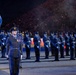 A formal military parade featuring personnel in Royal Air Force (RAF) dress uniforms performing at what appears to be a large nighttime ceremonial event, likely the Royal Edinburgh Military Tattoo or a similar prestigious military pageant. A senior officer stands at the forefront wearing multiple service medals, with a flag bearer displaying the RAF ensign (featuring the Union Jack and RAF roundel) to the left. Rows of uniformed RAF personnel stand at attention in the background before a large crowd of spectators.
