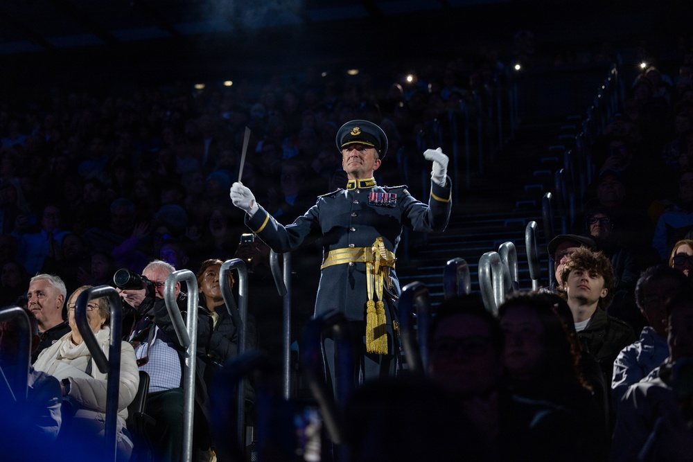 A military band conductor in formal dress uniform stands elevated among a large audience in what appears to be an indoor arena or stadium. He is actively conducting, wearing white gloves and holding a baton, with his arms raised in a conducting gesture. The scene is dramatically lit with the conductor as the focal point against a darkened crowd background.