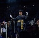 A military band conductor in formal dress uniform stands elevated among a large audience in what appears to be an indoor arena or stadium. He is actively conducting, wearing white gloves and holding a baton, with his arms raised in a conducting gesture. The scene is dramatically lit with the conductor as the focal point against a darkened crowd background.