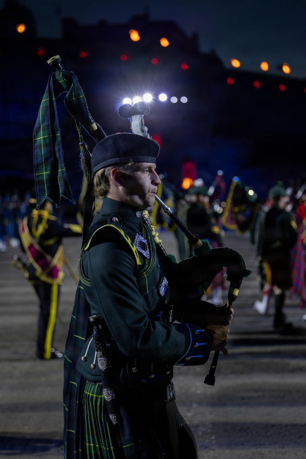 A bagpiper in dark green military dress uniform performs at what appears to be the Royal Edinburgh Military Tattoo, an annual outdoor performance event held at Edinburgh Castle. The performer is captured in sharp profile against a dramatically lit nighttime backdrop, with other band members and colorful stage lighting visible in the background. The dark green tartan kilt and uniform suggest affiliation with a Scottish or Irish military regiment.