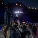 A bagpiper in dark green military dress uniform performs at what appears to be the Royal Edinburgh Military Tattoo, an annual outdoor performance event held at Edinburgh Castle. The performer is captured in sharp profile against a dramatically lit nighttime backdrop, with other band members and colorful stage lighting visible in the background. The dark green tartan kilt and uniform suggest affiliation with a Scottish or Irish military regiment.