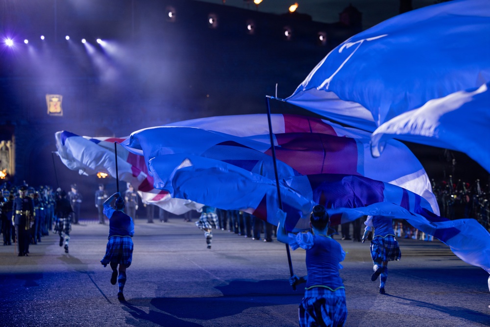 A nighttime ceremonial event featuring performers carrying large flags, including what appears to be the Scottish Saltire (blue with white diagonal cross) and the Union Jack. Participants are dressed in tartan/plaid attire, suggesting a Scottish military tattoo or similar formal parade event. The scene is dramatically lit with stage lighting creating a blue-toned atmosphere.