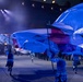 A nighttime ceremonial event featuring performers carrying large flags, including what appears to be the Scottish Saltire (blue with white diagonal cross) and the Union Jack. Participants are dressed in tartan/plaid attire, suggesting a Scottish military tattoo or similar formal parade event. The scene is dramatically lit with stage lighting creating a blue-toned atmosphere.