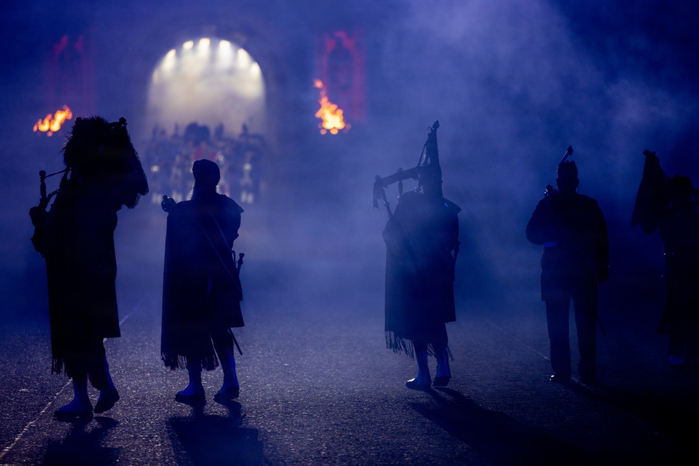 A dramatic nighttime scene showing silhouetted bagpipers and performers emerging through a smoke-filled archway with flames and stage lighting in the background. The atmospheric blue lighting and theatrical fog create a striking visual effect consistent with a large-scale military tattoo or ceremonial performance. The figures are dressed in traditional Scottish Highland attire including kilts and bearskin hats.