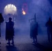 A dramatic nighttime scene showing silhouetted bagpipers and performers emerging through a smoke-filled archway with flames and stage lighting in the background. The atmospheric blue lighting and theatrical fog create a striking visual effect consistent with a large-scale military tattoo or ceremonial performance. The figures are dressed in traditional Scottish Highland attire including kilts and bearskin hats.