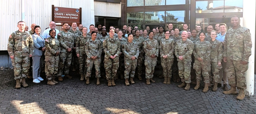 A large group of approximately 40-50 military personnel and a few civilians pose for a group photo outside the USAFE/AFAFRICA Conference Center. The majority are wearing OCP (Operational Camouflage Pattern) uniforms, suggesting a joint military gathering or conference. The setting appears to be a military installation, likely in Europe given the USAFE (United States Air Forces in Europe) signage.
