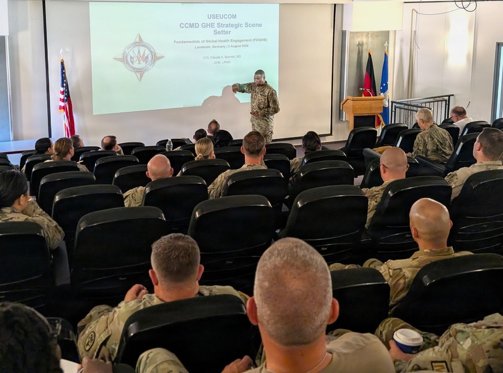 A military briefing is taking place in an auditorium-style room where a uniformed officer is presenting a USEUCOM CCMD GHE Strategic Scene Setter on Global Health Engagement (FOGHE). The presentation is dated 3 August 2020 and is located in Landstuhl, Germany, delivered by COL Claude A. Burnett, MD. The audience consists of approximately 20-30 military personnel in camouflage uniforms seated in black chairs.