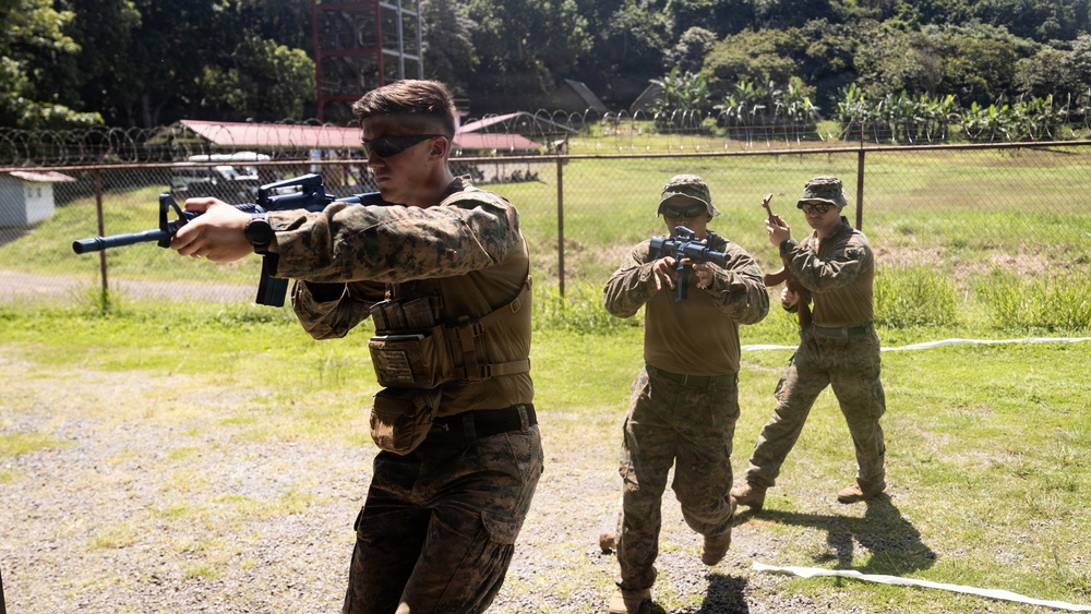 Three military personnel in camouflage uniforms are conducting a weapons training exercise at an outdoor range or training facility. The foreground soldier is aiming an M4-style rifle while two others in the background appear to be observing or participating in the drill. The setting includes a fenced perimeter with barbed wire, lush green vegetation, and a red-roofed structure in the background.