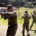 Three military personnel in camouflage uniforms are conducting a weapons training exercise at an outdoor range or training facility. The foreground soldier is aiming an M4-style rifle while two others in the background appear to be observing or participating in the drill. The setting includes a fenced perimeter with barbed wire, lush green vegetation, and a red-roofed structure in the background.