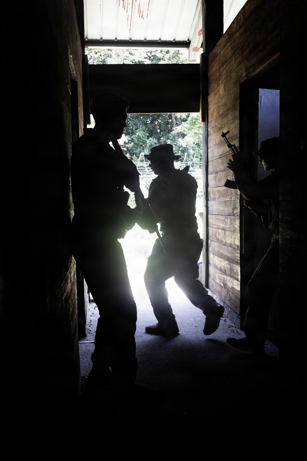 The image shows silhouetted figures armed with long firearms moving through a narrow corridor or doorway of a wooden structure. The strong backlight from an outdoor area creates dramatic contrast, rendering the figures almost entirely in shadow. The scene appears to depict a tactical entry or clearing operation, possibly military or law enforcement training.