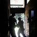 The image shows silhouetted figures armed with long firearms moving through a narrow corridor or doorway of a wooden structure. The strong backlight from an outdoor area creates dramatic contrast, rendering the figures almost entirely in shadow. The scene appears to depict a tactical entry or clearing operation, possibly military or law enforcement training.