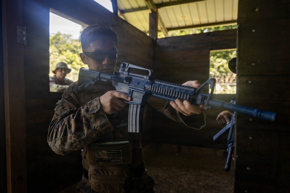 A U.S. Marine in MARPAT camouflage uniform is aiming an M16-style rifle inside a wooden training structure. A second military personnel member is visible in the background observing or participating in the exercise. The setting appears to be a military training range or shoot house.