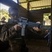 A U.S. Marine in MARPAT camouflage uniform is aiming an M16-style rifle inside a wooden training structure. A second military personnel member is visible in the background observing or participating in the exercise. The setting appears to be a military training range or shoot house.
