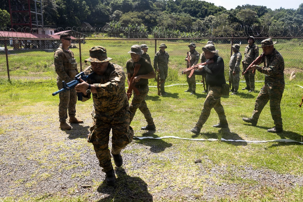 A military training exercise is taking place outdoors at what appears to be a secured compound or base. Multiple soldiers in camouflage uniforms are practicing weapons handling and tactical drills, with at least one instructor observing. The training involves both blue-barreled training rifles and standard-colored rifles, indicating a live-fire simulation or force-on-force exercise.