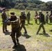 A military training exercise is taking place outdoors at what appears to be a secured compound or base. Multiple soldiers in camouflage uniforms are practicing weapons handling and tactical drills, with at least one instructor observing. The training involves both blue-barreled training rifles and standard-colored rifles, indicating a live-fire simulation or force-on-force exercise.