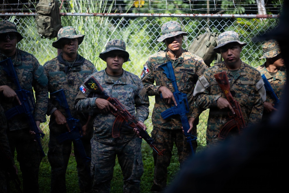 A group of military personnel in digital camouflage uniforms and boonie hats are standing together, holding rifles with blue training barrel attachments, indicating a training exercise. At least one soldier displays a Panamanian flag patch on their sleeve, suggesting this involves Panamanian security forces. The group appears to be receiving instruction or a briefing near a chain-link fence in a tropical/jungle environment.