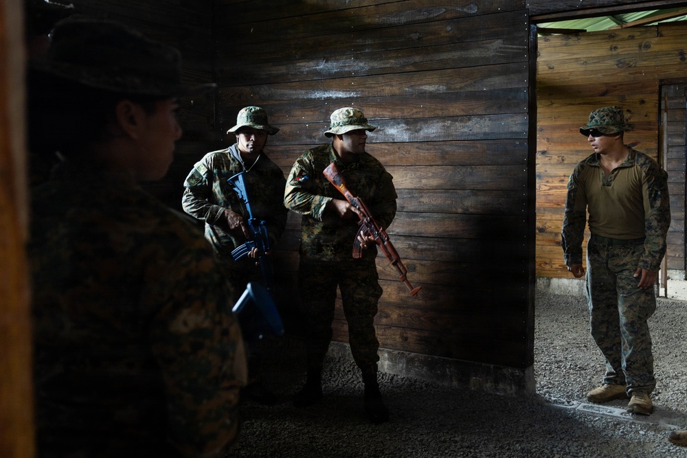 The image shows a group of military personnel inside a wooden structure, appearing to be engaged in a training exercise. Three soldiers in camouflage uniforms and bush hats are holding rifles with colored (blue and orange) training weapon indicators, while a fourth individual in a different camouflage pattern stands near a doorway, likely in an instructor or observer role. The setting and colored training weapons suggest this is a military training or instruction scenario.