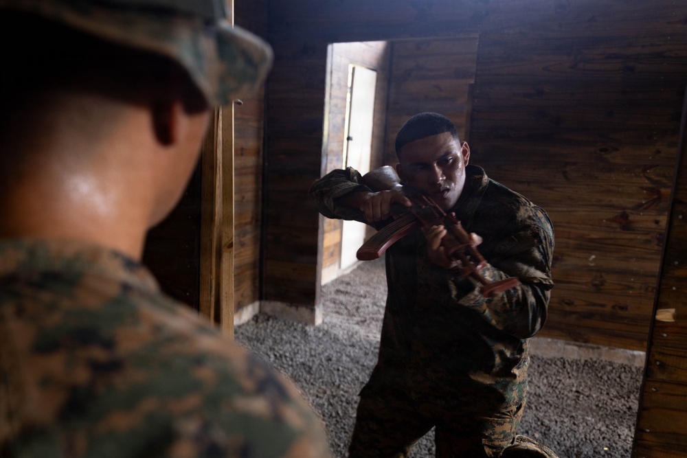 Two individuals in U.S. Marine Corps MARPAT camouflage uniforms are inside a wooden structure, appearing to conduct close-quarters combat or martial arts training. The foreground Marine is partially visible from behind, while the background Marine is in an aggressive stance holding what appears to be a training weapon or rubber knife. The setting appears to be a military training facility with a gravel floor and wooden walls.