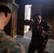 Two individuals in U.S. Marine Corps MARPAT camouflage uniforms are inside a wooden structure, appearing to conduct close-quarters combat or martial arts training. The foreground Marine is partially visible from behind, while the background Marine is in an aggressive stance holding what appears to be a training weapon or rubber knife. The setting appears to be a military training facility with a gravel floor and wooden walls.