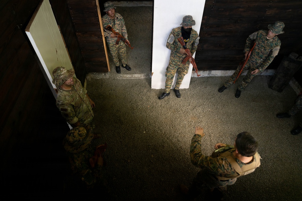 An overhead/elevated view of a military training exercise taking place inside a building or shoot house. A instructor or trainer appears to be briefing or directing several soldiers wearing mixed camouflage uniforms. The personnel are holding training rifles (appearing to be red/orange-colored simunition or training weapons) and are positioned near doorways, suggesting room-clearing or close-quarters battle training.