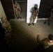 An overhead/elevated view of a military training exercise taking place inside a building or shoot house. A instructor or trainer appears to be briefing or directing several soldiers wearing mixed camouflage uniforms. The personnel are holding training rifles (appearing to be red/orange-colored simunition or training weapons) and are positioned near doorways, suggesting room-clearing or close-quarters battle training.