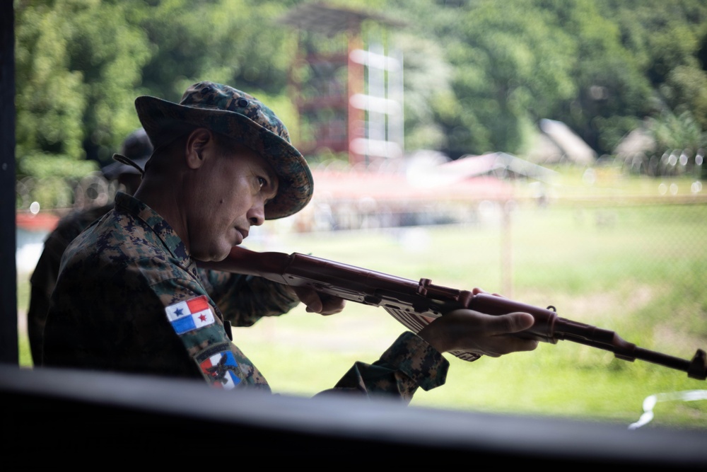 A soldier in digital camouflage uniform and boonie hat aims a rifle at what appears to be a shooting range. The soldier wears a patch on the left sleeve depicting the flag of Panama, identifying the national affiliation. The setting appears to be an outdoor firing range with green vegetation and structures visible in the background.