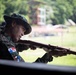 A soldier in digital camouflage uniform and boonie hat aims a rifle at what appears to be a shooting range. The soldier wears a patch on the left sleeve depicting the flag of Panama, identifying the national affiliation. The setting appears to be an outdoor firing range with green vegetation and structures visible in the background.