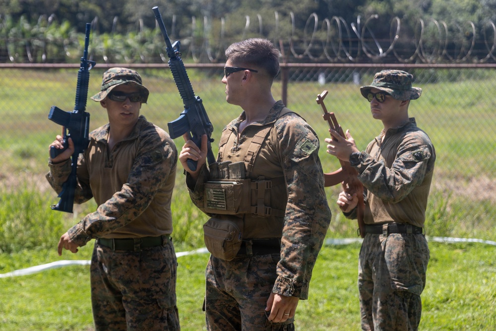 Three military personnel in MARPAT (Marine pattern) camouflage uniforms are standing on a grassy field holding rifles in an upright, safe carry position. The setting appears to be a military training range or base, evidenced by the razor wire fence visible in the background. The individuals appear to be U.S. Marines engaged in a weapons handling drill or training exercise.