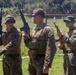 Three military personnel in MARPAT (Marine pattern) camouflage uniforms are standing on a grassy field holding rifles in an upright, safe carry position. The setting appears to be a military training range or base, evidenced by the razor wire fence visible in the background. The individuals appear to be U.S. Marines engaged in a weapons handling drill or training exercise.