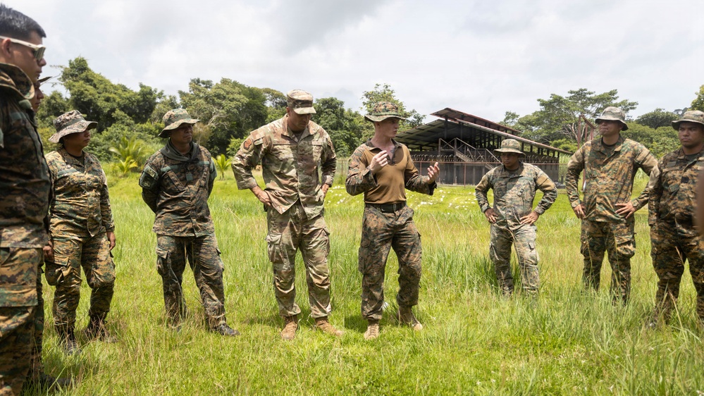 A group of military personnel in various camouflage uniforms are gathered in an outdoor grassy field, appearing to be in a briefing or instructional session. One individual in a tan/coyote brown shirt is speaking and gesturing to the group, suggesting a leadership or instructor role. The setting appears to be a tropical training environment, with lush vegetation and a covered structure visible in the background.