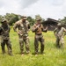 A group of military personnel in various camouflage uniforms are gathered in an outdoor grassy field, appearing to be in a briefing or instructional session. One individual in a tan/coyote brown shirt is speaking and gesturing to the group, suggesting a leadership or instructor role. The setting appears to be a tropical training environment, with lush vegetation and a covered structure visible in the background.