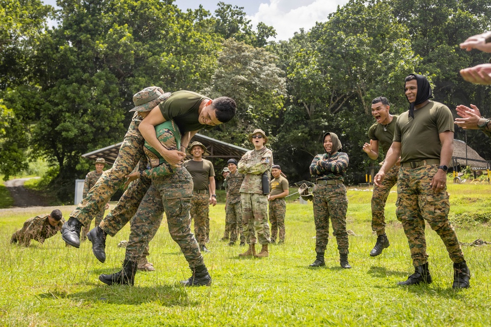 A group of military personnel in various camouflage uniforms are gathered on a grassy field during what appears to be a training exercise or team-building activity. Two soldiers in the foreground are engaged in a grappling or combat training demonstration, with one lifting the other off the ground. Onlookers in the background are reacting with laughter and applause, suggesting a lighthearted or competitive training environment.