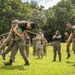 A group of military personnel in various camouflage uniforms are gathered on a grassy field during what appears to be a training exercise or team-building activity. Two soldiers in the foreground are engaged in a grappling or combat training demonstration, with one lifting the other off the ground. Onlookers in the background are reacting with laughter and applause, suggesting a lighthearted or competitive training environment.