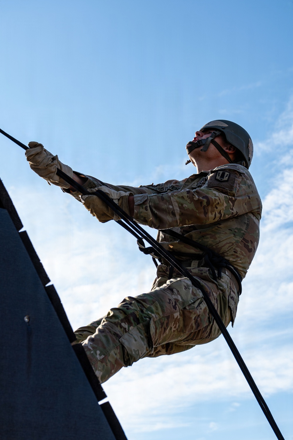 A military service member in MultiCam/OCP camouflage uniform is rappelling down a vertical structure during what appears to be a training exercise. The photo is taken from a low angle looking upward, with a blue sky as the backdrop. The soldier is equipped with a helmet, protective eyewear, gloves, and a rappelling harness with black ropes.