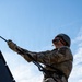 A military service member in MultiCam/OCP camouflage uniform is rappelling down a vertical structure during what appears to be a training exercise. The photo is taken from a low angle looking upward, with a blue sky as the backdrop. The soldier is equipped with a helmet, protective eyewear, gloves, and a rappelling harness with black ropes.