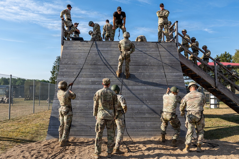 Military personnel are conducting rappelling training on a wooden tower structure at what appears to be a military training facility. One soldier is actively rappelling down the face of the wooden wall while instructors and other trainees observe from the top platform and ground level. The exercise involves rope management with multiple soldiers on the ground acting as belayers or waiting their turn.
