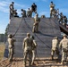 Military personnel are conducting rappelling training on a wooden tower structure at what appears to be a military training facility. One soldier is actively rappelling down the face of the wooden wall while instructors and other trainees observe from the top platform and ground level. The exercise involves rope management with multiple soldiers on the ground acting as belayers or waiting their turn.