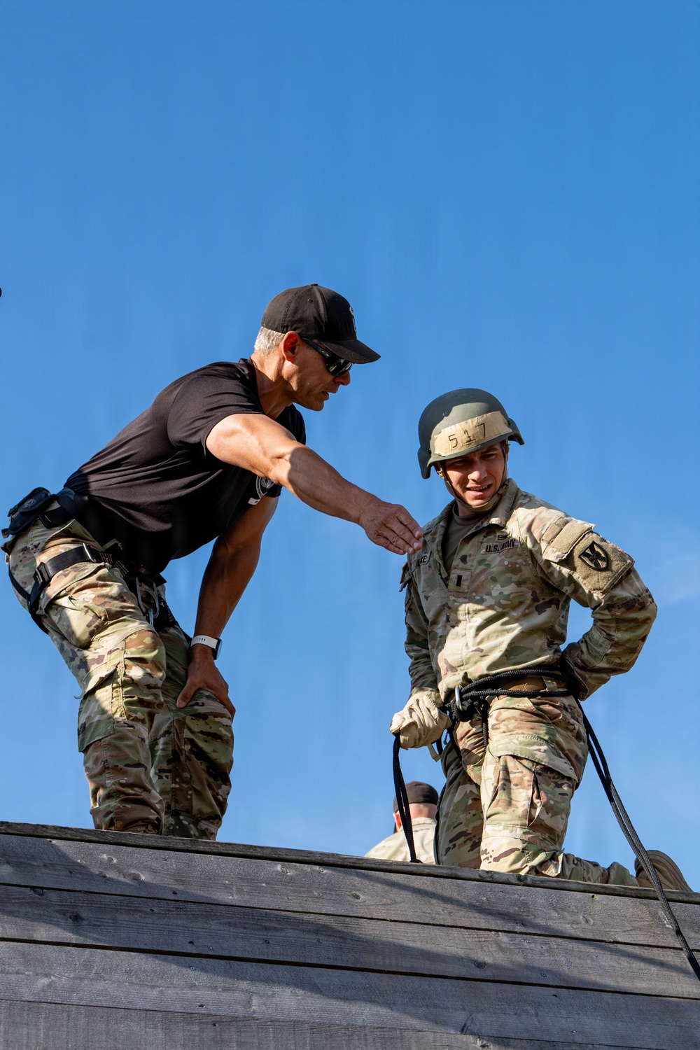 Two individuals are positioned at the top of a wooden structure, likely a rappelling or obstacle course tower. An instructor in a black t-shirt and camouflage pants is leaning toward a U.S. Army soldier wearing a helmet marked '517' and full OCP (Operational Camouflage Pattern) uniform with rappelling harness and rope. The interaction appears to be instructional guidance prior to a rappel descent.